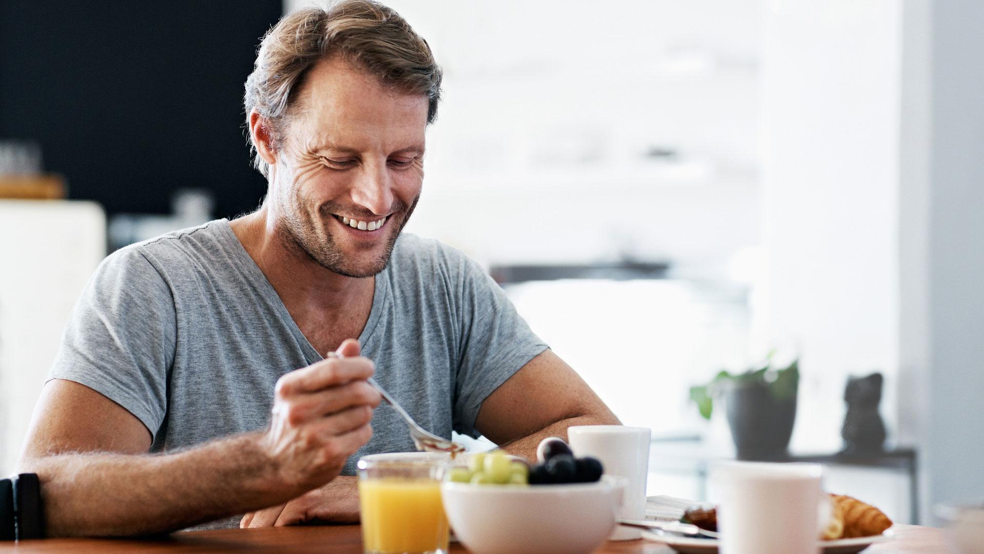 En mand sidder ved et bord og smiler mens han spiser morgenmad.