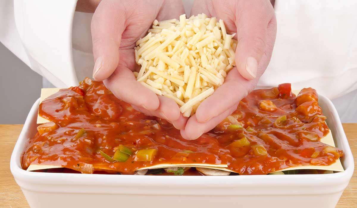 En håndfuld revet ost drysses over en lasagne.