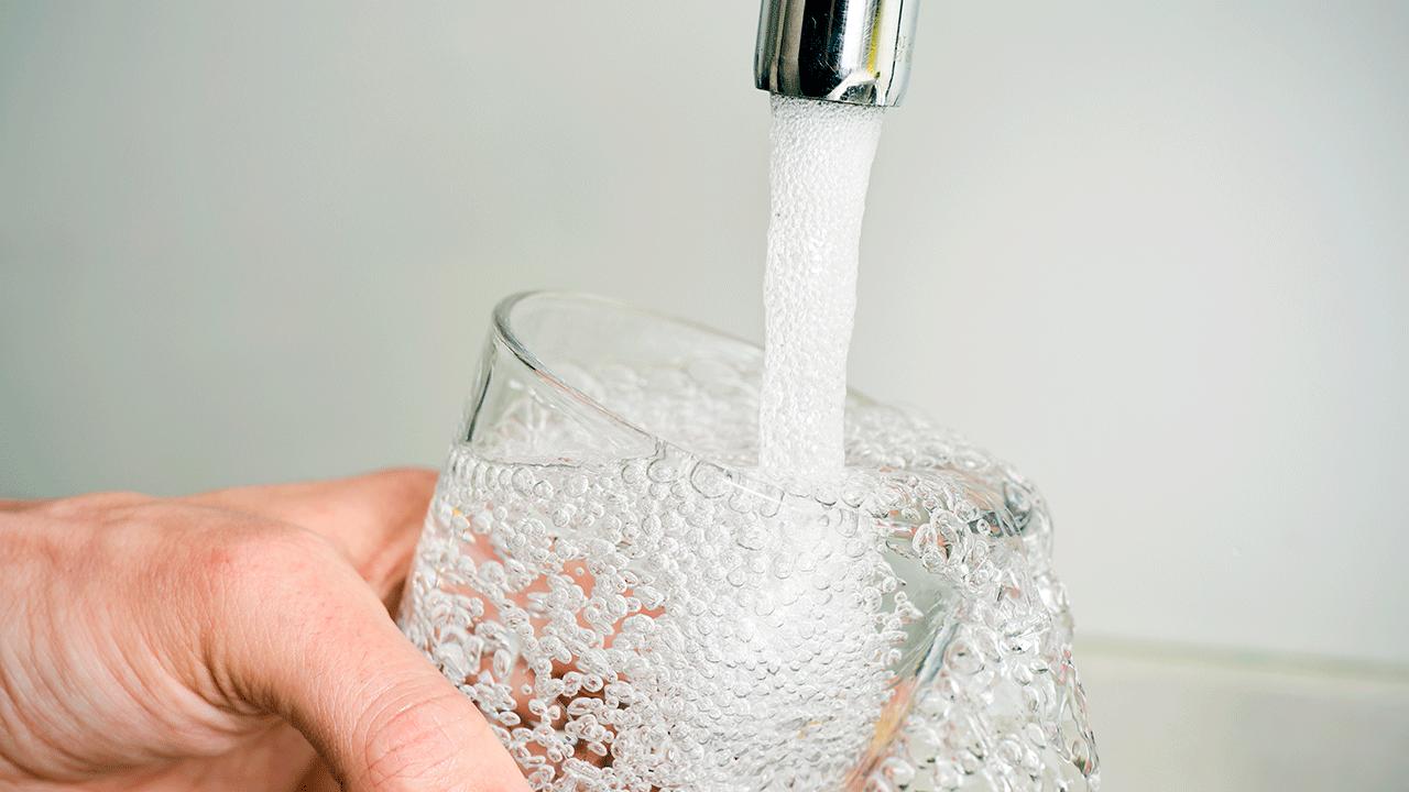 Hånd holder glas, der fyldes med vand fra vandhane på lys baggrund. 