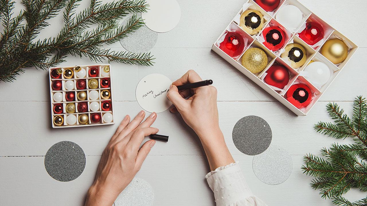 Foto set oppefra af hænder, der skriver på til-og frakort mellem julekugler og gran.