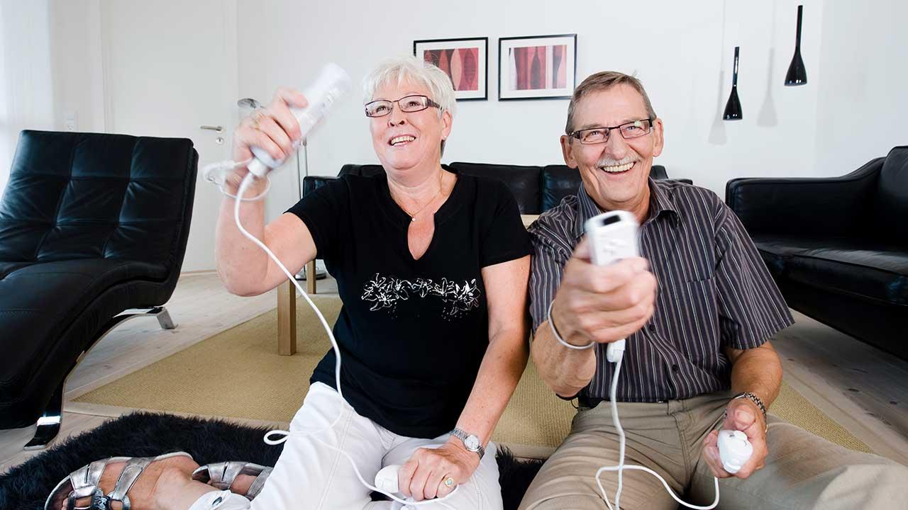 Et ældre ægtepar sidder på gulvet i deres stue med controllere i hånden og spiller computerspil mens de griner og har det sjovt. 
