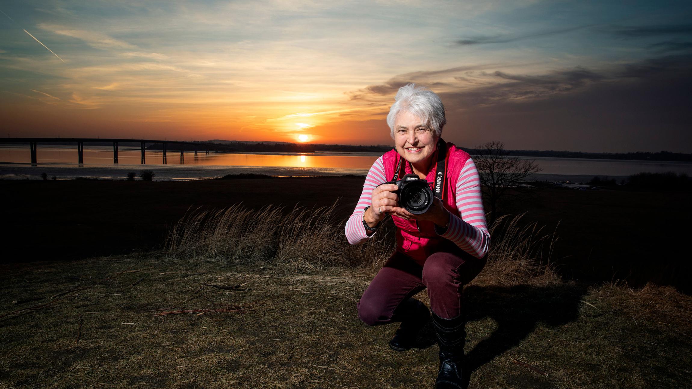 Kvinde på hug med kamera i hånden foran solnedgang