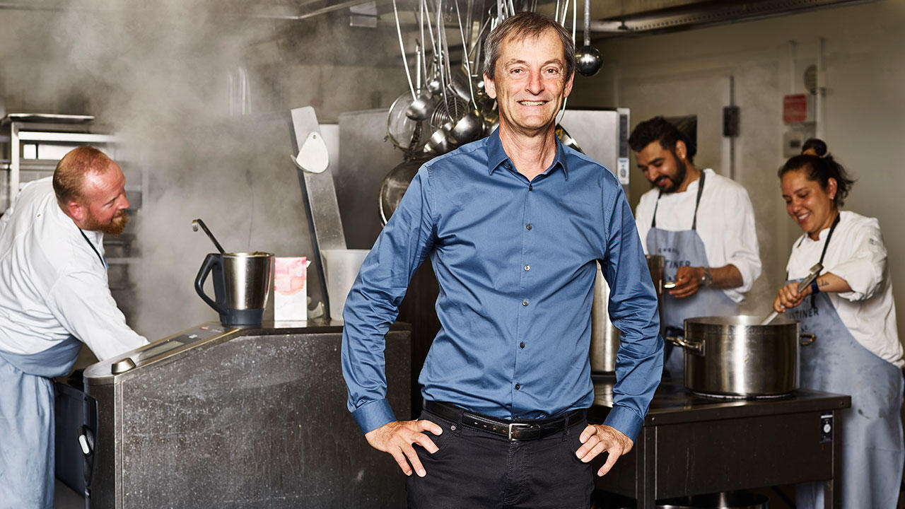 Claus Tingstrøm står i kantine foran kokke der arbejder og laver mad